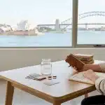 Eine Frau an einem Holztisch mit einem Lederjournal in der Hand, die aus dem Fenster auf die Skyline von Sydney blickt.